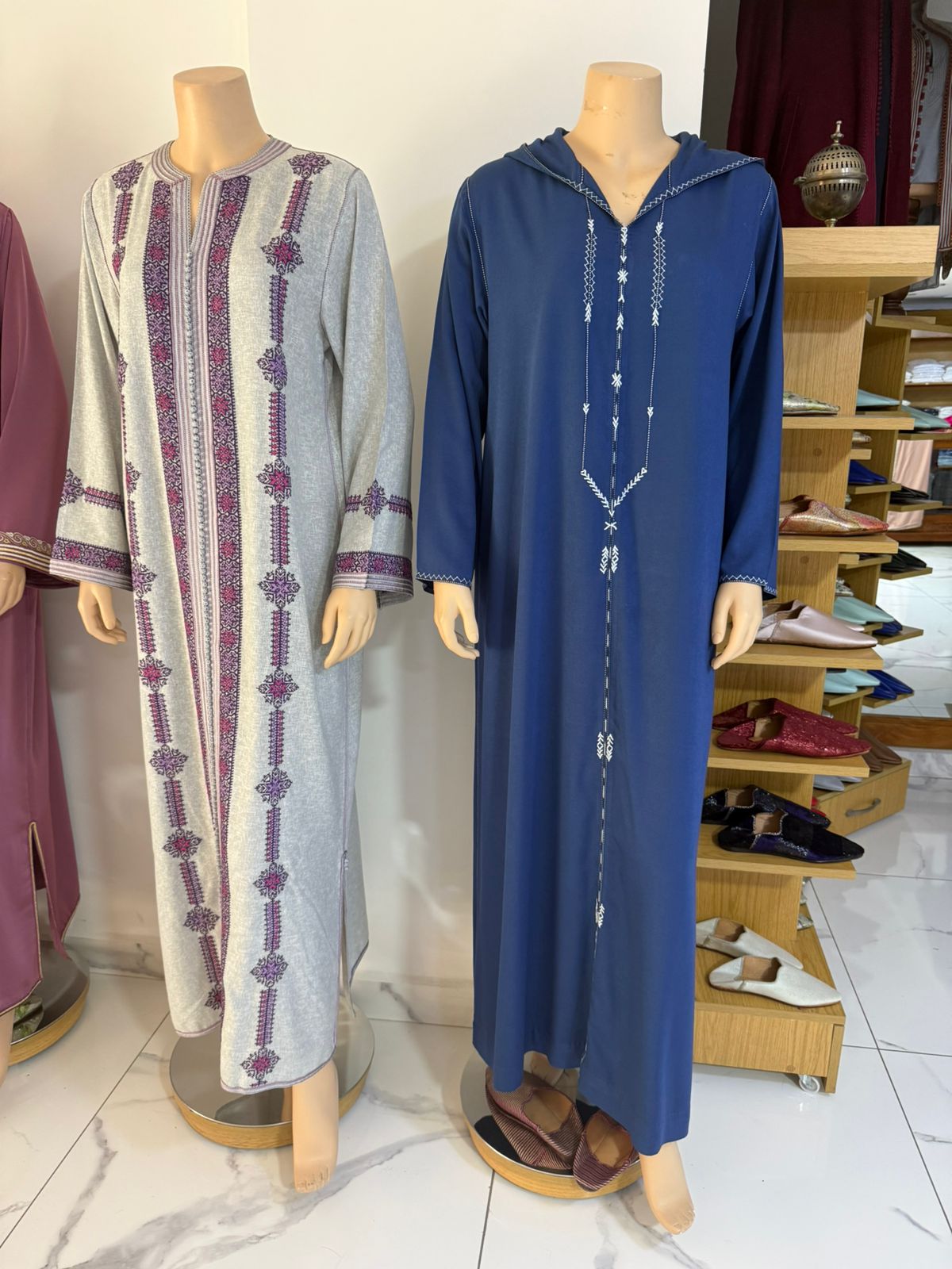 Djellaba femme à capuche bleu indigo, terz blanc ton sur ton en chevrons sur le plastron, présentée à côté d'une autre djellaba femme grise à broderie prune, Tazi Caftan Marrakech